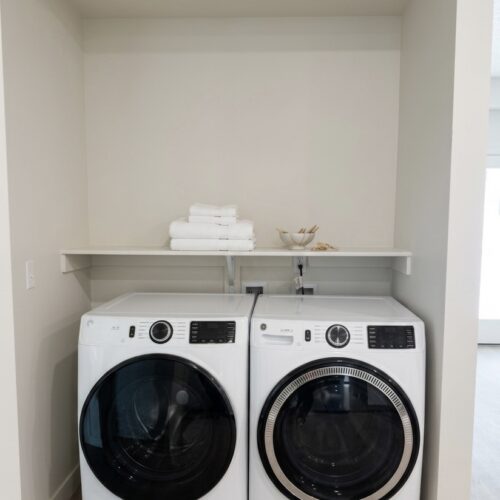 White front-loading washer and dryer side by side in a small laundry alcove with a shelf above and folded towels on the shelf