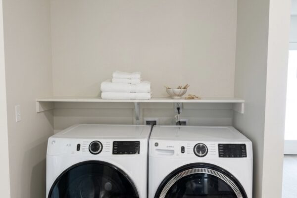 White front-loading washer and dryer side by side in a small laundry alcove with a shelf above and folded towels on the shelf
