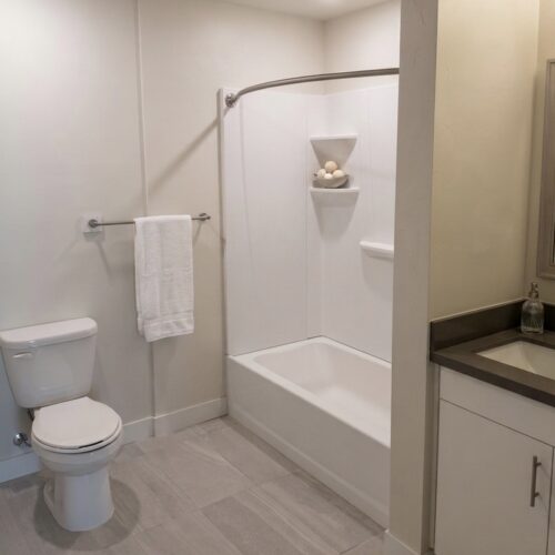 Bright, modern bathroom featuring a toilet on the left, towel bar with a white towel, and a white tub/shower with a curved rod and built-in corner shelves; light gray floor tiles and a vanity with dark countertop on the right.