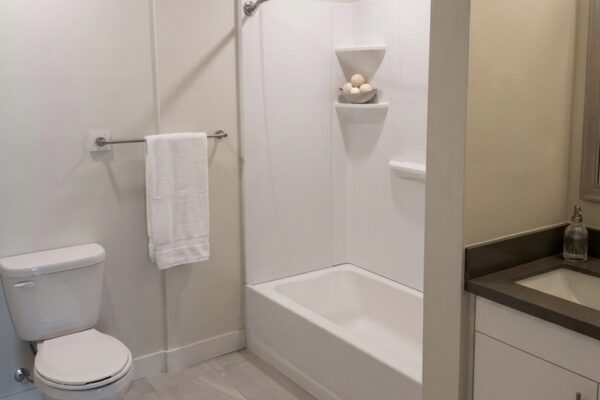Bright, modern bathroom featuring a toilet on the left, towel bar with a white towel, and a white tub/shower with a curved rod and built-in corner shelves; light gray floor tiles and a vanity with dark countertop on the right.