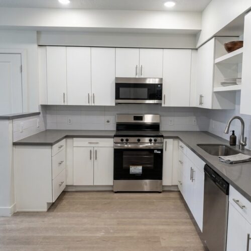 Modern L-shaped kitchen with white cabinets, gray countertops, and stainless steel appliances (oven, microwave, dishwasher)