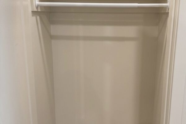 Narrow empty closet with a white clothes rod and a top shelf, beige walls, and light wood floor.