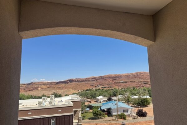 View of a desert town framed by an arched patio opening, with red rock hills under a blue sky.