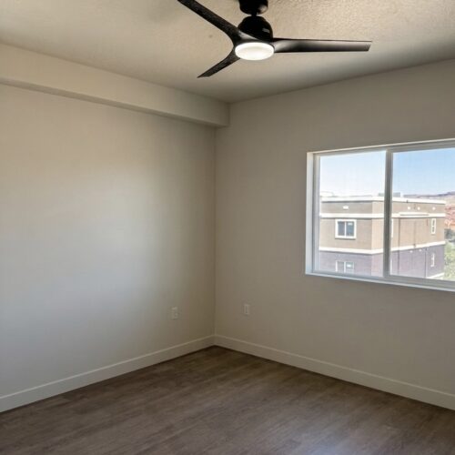 Empty bedroom with a black three-blade ceiling fan, white walls, wood-look flooring, and a window showing a neighboring building outside.