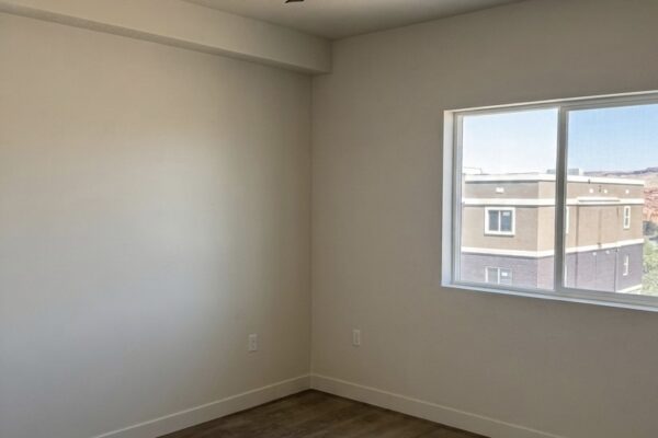 Empty bedroom with a black three-blade ceiling fan, white walls, wood-look flooring, and a window showing a neighboring building outside.